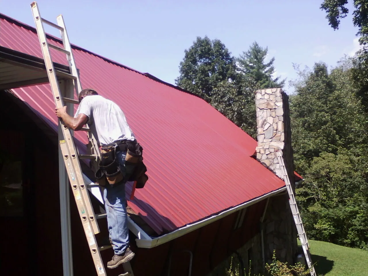 Roofer on a ladder beside a red metal roof and stone chimney — Soffit & Fascia Repair by Roofing Brothers in Mccloud
